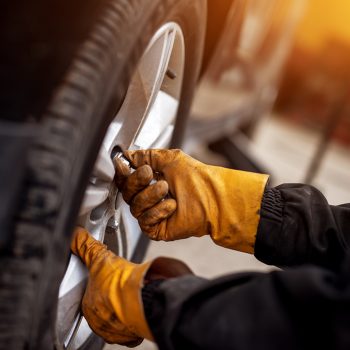An experienced mechanic with orange gloves is putting screws on a placed wheel on a car.