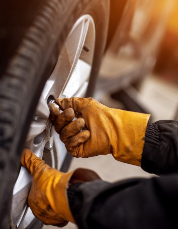 An experienced mechanic with orange gloves is putting screws on a placed wheel on a car.